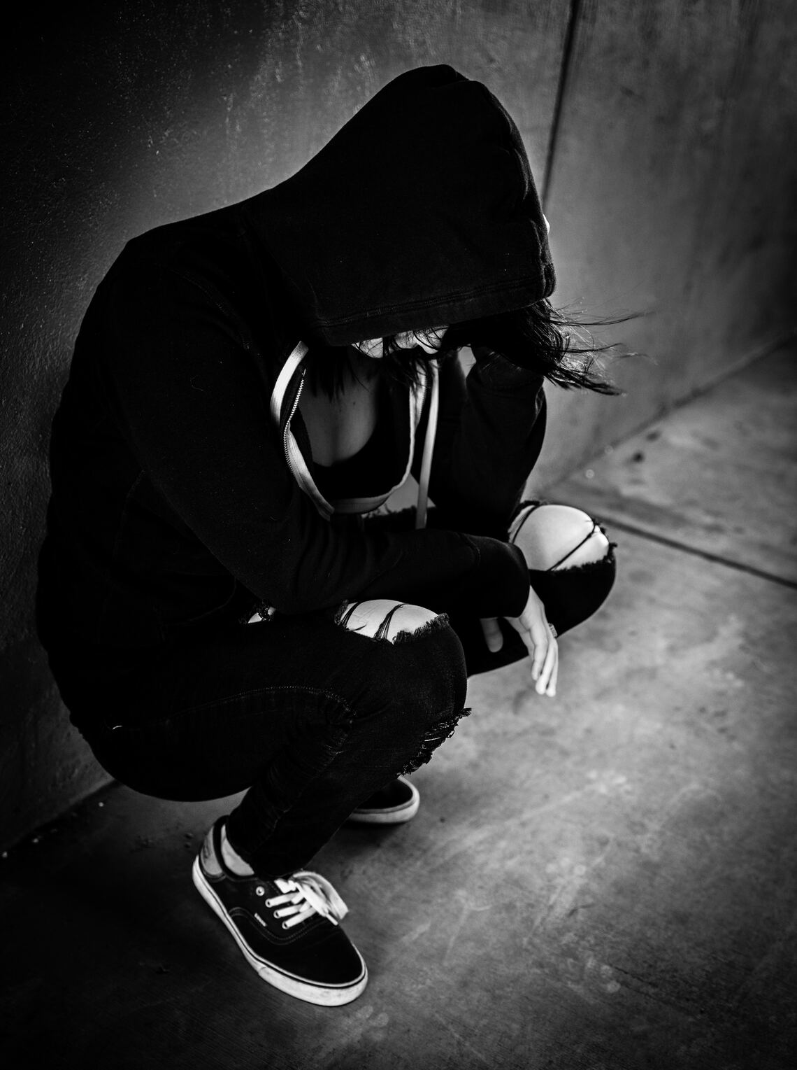 A young woman in a hoodie with her head down leaning against a wall